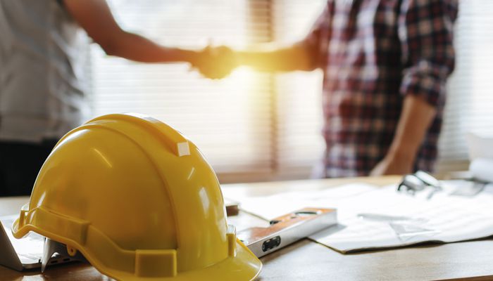Two people shaking hands behind a table with glasses, paper, a leveler, and a yellow hat on top